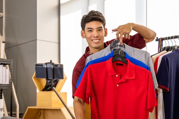 Joven bloguero comerciante en línea masculino asiático que usa una cámara de teléfono móvil con selfie stick para video en vivo para revisar y vender productos mirando la cámara Influenciador de las redes sociales y concepto de compras en línea