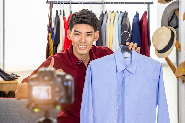 Joven bloguero comerciante en línea masculino asiático que usa cámara para video en vivo para revisar y vender productos con el pulgar hacia arriba Influenciador de medios sociales y concepto de compras en línea
