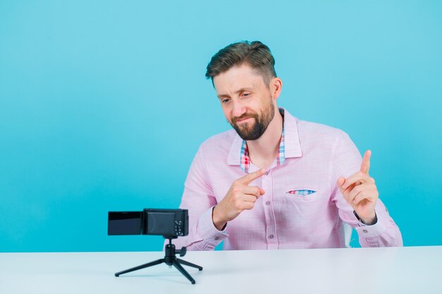 El joven bloguero está posando para su mini cámara apuntando a la izquierda con los dedos índices sobre fondo azul.