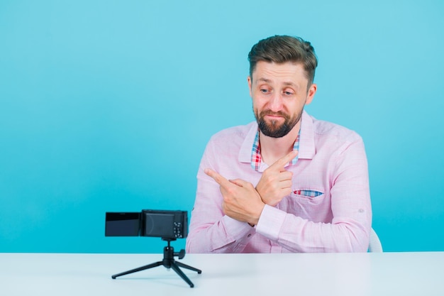 El joven bloguero está posando para su mini cámara cruzando las manos sobre fondo azul.