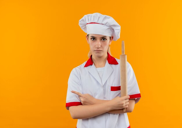 Joven bonita cocinera en uniforme de chef sosteniendo un rodillo y apuntando a un lado