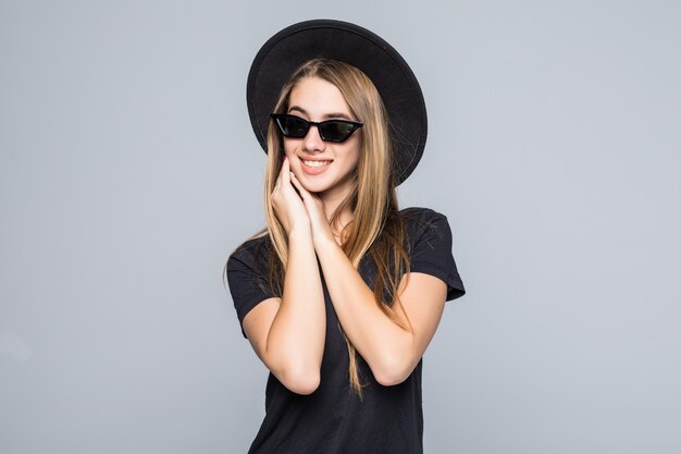 Joven bonita a dama sonriente con gafas de sol brillantes vestida con sombrero negro, camiseta negra y pantalón oscuro aislado sobre fondo gris tiene las manos debajo de la barbilla juntas