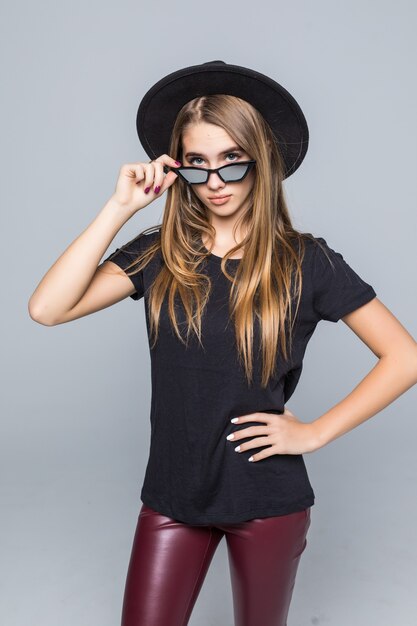 Joven bonita a dama sonriente con gafas de sol brillantes vestida con sombrero negro, camiseta negra y pantalón oscuro aislado sobre fondo gris