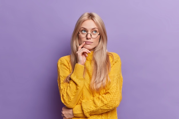 Joven bonita a joven piensa en ideas concentradas por encima de pie pensativo y mantiene la mano en la cara se encuentra en pose pensativa lleva gafas redondas suéter amarillo