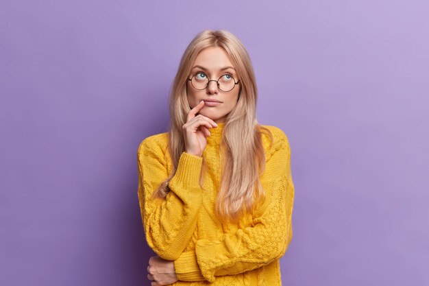 Joven bonita a joven piensa en ideas concentradas por encima de pie pensativo y mantiene la mano en la cara se encuentra en pose pensativa lleva gafas redondas suéter amarillo