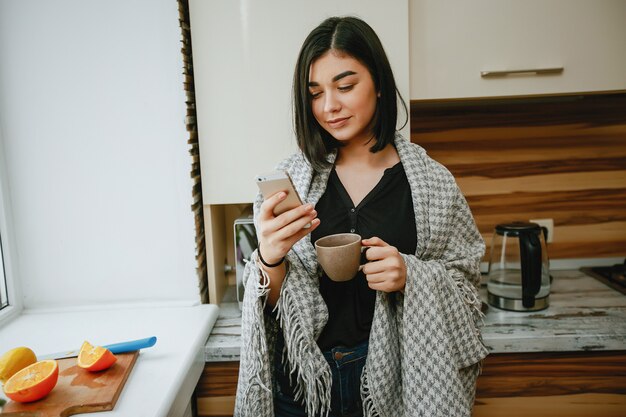 joven y bonita morena de pie en la cocina con teléfono y bebiendo un café