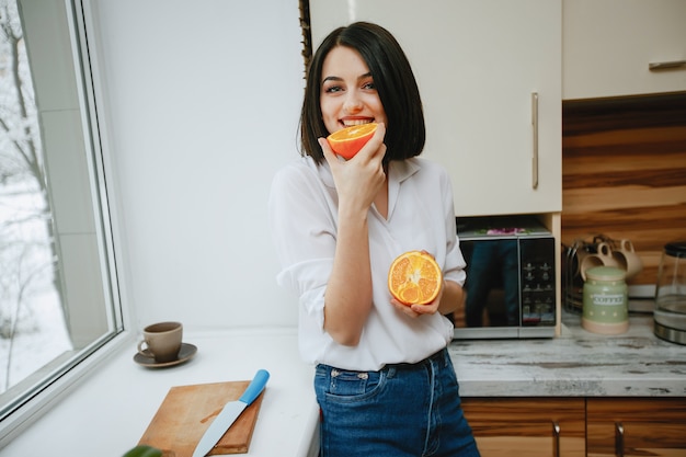 joven y bonita morena de pie junto a la ventana en la cocina con naranja