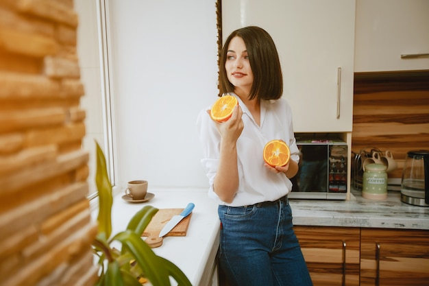 joven y bonita morena de pie junto a la ventana en la cocina con naranja