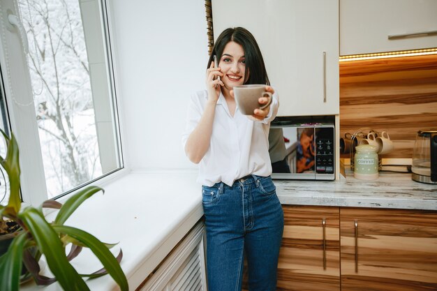 joven y bonita morena de pie junto a la ventana en la cocina con teléfono y café