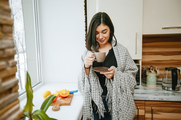 joven y bonita morena de pie junto a la ventana en la cocina y tomando un café