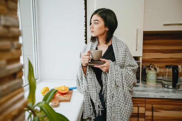 joven y bonita morena de pie junto a la ventana en la cocina y tomando un café