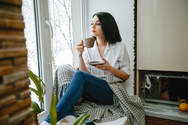 joven y bonita morena sentada junto a la ventana en la cocina con café