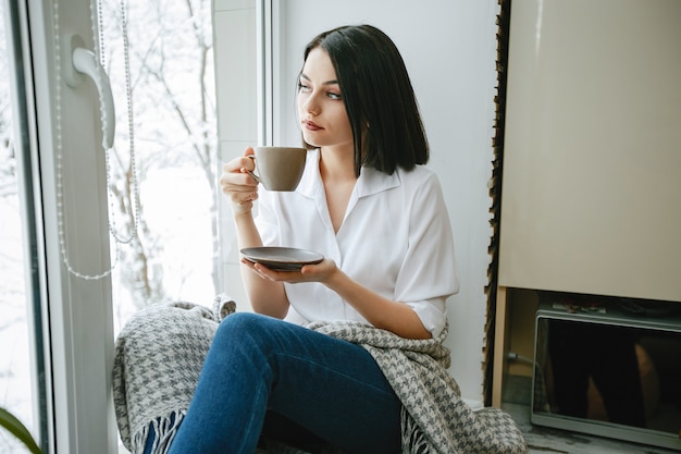 joven y bonita morena sentada junto a la ventana en la cocina con café