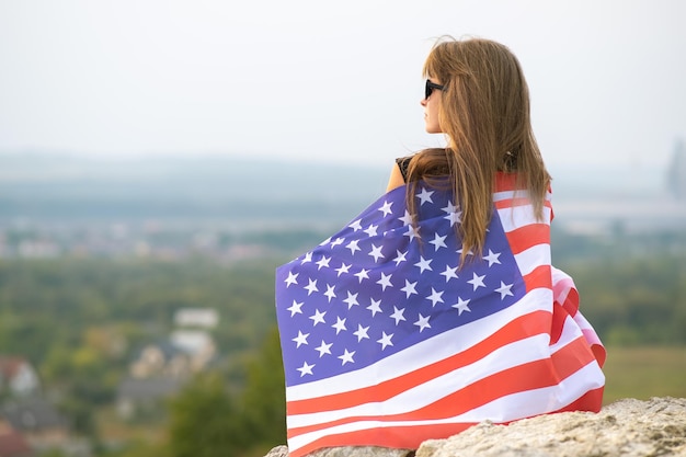 Joven bonita a mujer americana con pelo largo sosteniendo ondeando en el viento la bandera de estados unidos en sus hombros descansando al aire libre disfrutando de un cálido día de verano.