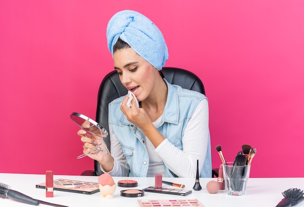 Joven bonita a mujer caucásica con el pelo envuelto en una toalla sentada en la mesa con herramientas de maquillaje sosteniendo y mirando el espejo secándose la boca con una servilleta aislada en la pared rosa con espacio de copia