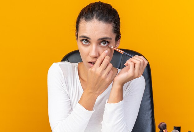 Joven bonita a mujer caucásica sentada a la mesa con herramientas de maquillaje aplicando sombra de ojos con pincel de maquillaje aislado en la pared naranja con espacio de copia