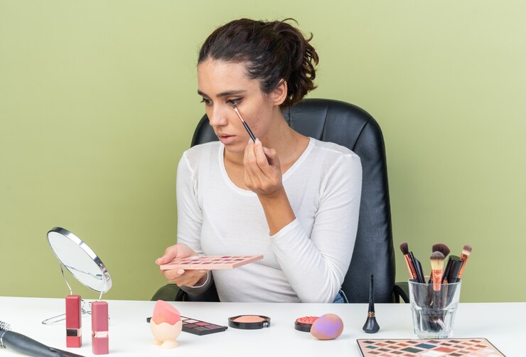 Foto gratuito joven bonita a mujer caucásica sentada a la mesa con herramientas de maquillaje aplicando sombra de ojos y sosteniendo la paleta de sombra de ojos aislada en la pared verde oliva con espacio de copia