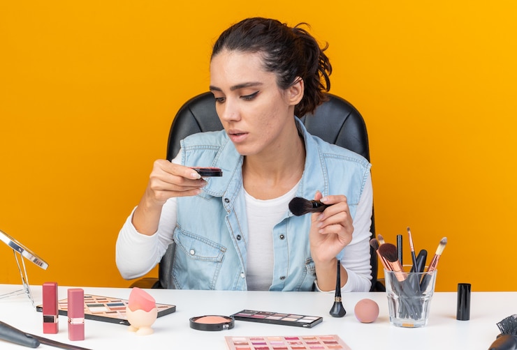 Foto gratuito joven bonita a mujer caucásica sentada a la mesa con herramientas de maquillaje sosteniendo pincel de maquillaje y mirando rubor