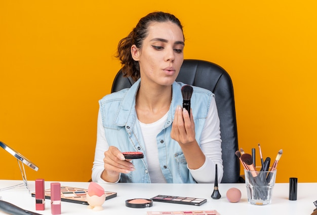 Joven bonita a mujer caucásica sentada a la mesa con herramientas de maquillaje sosteniendo rubor y soplando en pincel de maquillaje