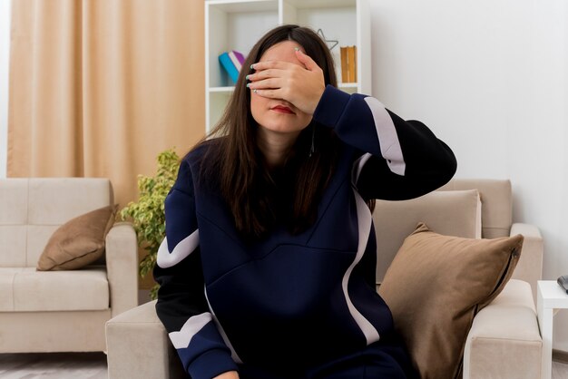 Joven bonita a mujer caucásica sentada en un sillón en la sala de estar diseñada cubriendo los ojos con la mano