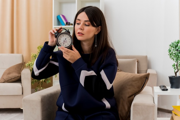 Joven bonita a mujer caucásica sentada en un sillón en la sala de estar diseñada sosteniendo y mirando el despertador