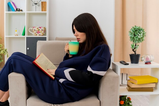 Joven bonita a mujer caucásica sentada en un sillón en la sala de estar diseñada sosteniendo una taza con el libro en las piernas tocando y leyendo el libro y preparándose para tomar café