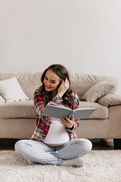 Joven bonita a mujer embarazada en camisa a cuadros rojos y pantalones de mezclilla leyendo un libro y sonriendo