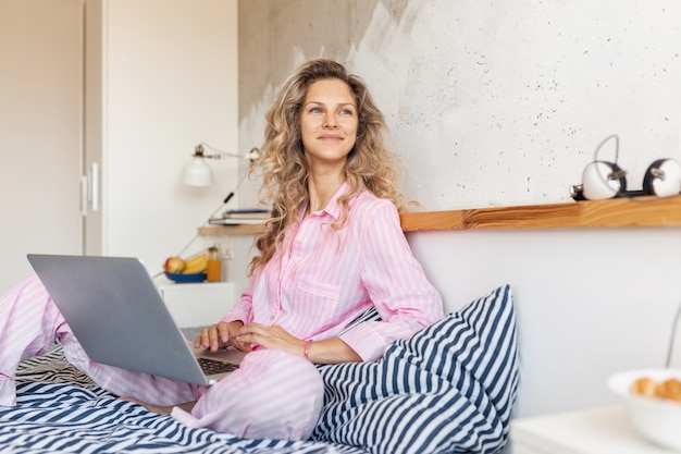 Joven bonita a mujer rubia en pijama rosa sentada en la cama trabajando en la computadora portátil