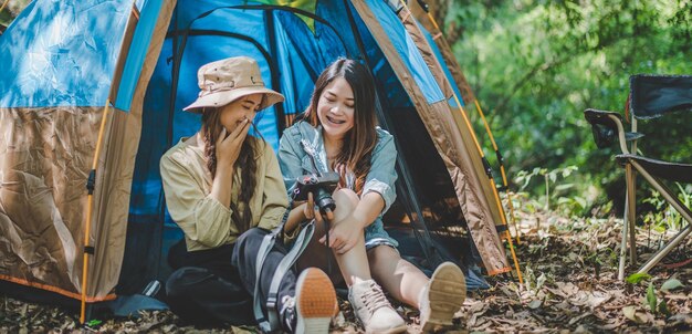 Joven bonita tomando una foto con la cámara y mostrándole una foto a su amiga mientras está sentada en la tienda de campaña en el bosque Mujeres jóvenes del grupo asiático viajan de campamento al aire libre