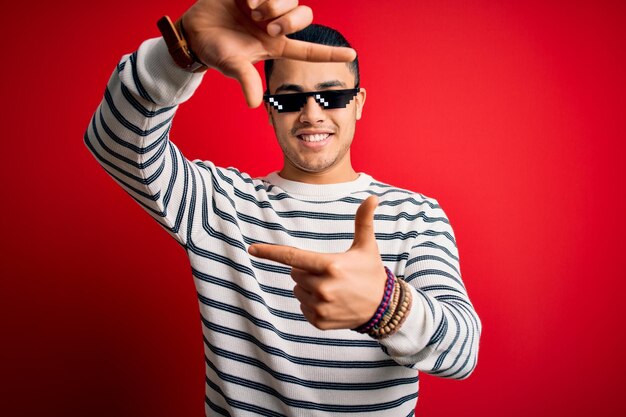 Joven brasileño con gafas de sol divertidas de thug life sobre fondo rojo aislado sonriendo haciendo marco con las manos y los dedos con cara feliz Creatividad y concepto de fotografía