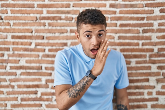 Foto gratuita joven brasileño parado sobre una pared de ladrillo con la mano en la boca contando rumores secretos susurrando una conversación maliciosa