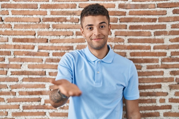 Foto gratuita joven brasileño parado sobre una pared de ladrillo sonriendo alegre ofreciendo la mano de la palma dando asistencia y aceptación