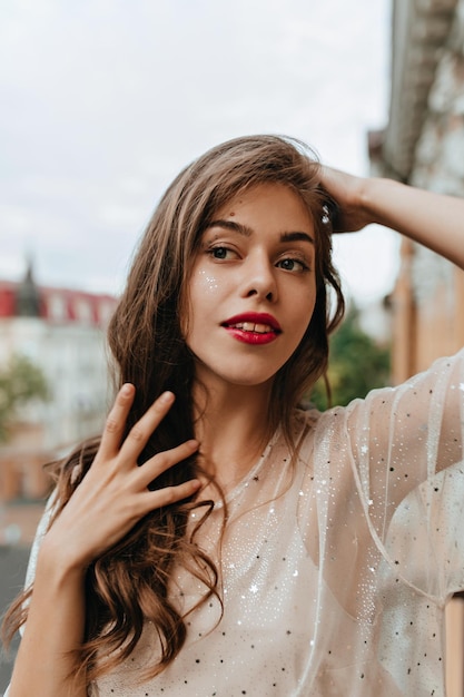 Joven bronceada con labios rojos y destellos en la cara posa afuera Chica atractiva con vestido blanco toca el cabello y posa en el balcón