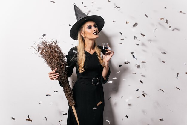 Joven bruja alegre bebiendo vino. Mujer despreocupada en traje de halloween posando en la fiesta.