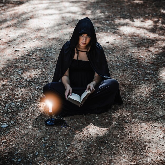 Joven bruja con el libro y el candelero ardiente en el suelo del bosque