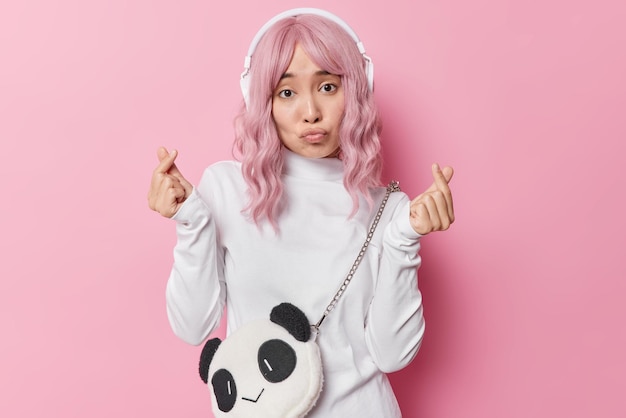 Una joven de cabello bastante rosado hace un gesto de mini corazón expresa amor mantiene los labios redondeados usa un cuello de tortuga blanco escucha música a través de auriculares aislados sobre un fondo rosado. Coreano como signo.