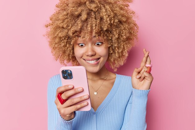 Una joven de cabello rizado cruza los dedos para tener buena suerte, se ve en la pantalla del teléfono inteligente sentado, espera los resultados, usa poses casuales de puente azul sobre fondo rosa, cree en la buena fortuna y espera ganar la lotería.