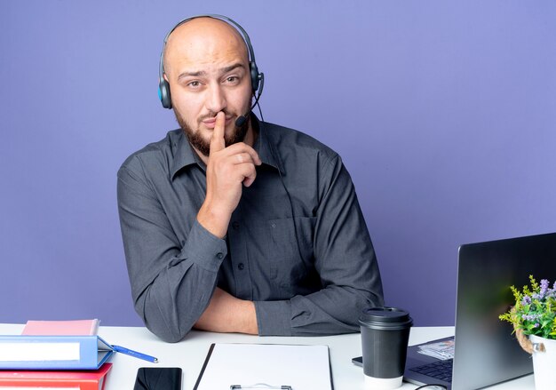 Joven calvo call center hombre con auriculares sentado en un escritorio con herramientas de trabajo gesticulando silencio aislado sobre fondo púrpura