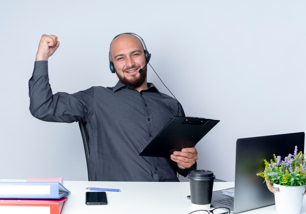 Joven calvo call center hombre con auriculares sentado en un escritorio con herramientas de trabajo sosteniendo el portapapeles aislado sobre fondo blanco.