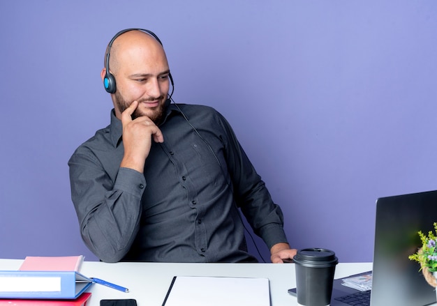 Joven calvo call center hombre con auriculares sentados en un escritorio con herramientas de trabajo aisladas sobre fondo púrpura