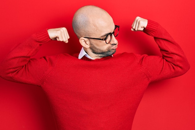 Joven calvo con ropa informal y gafas mostrando los músculos de los brazos sonriendo orgulloso. concepto de fitness.