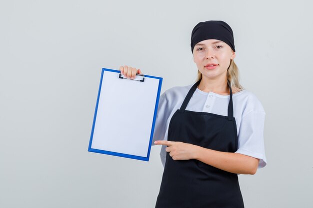 Joven camarera apuntando al portapapeles en uniforme y delantal