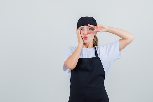 Foto gratuita joven camarera mostrando v-sign con labios doblados en uniforme y delantal