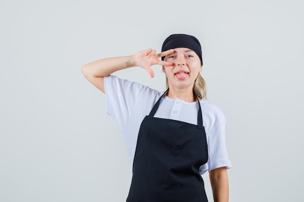 Joven camarera que muestra el signo v cerca del ojo en uniforme y delantal y parece feliz