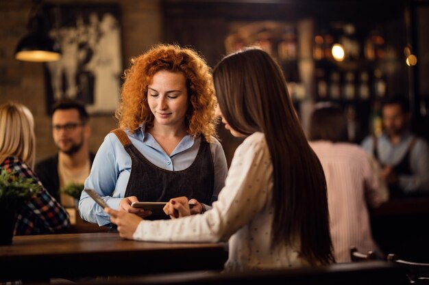 Joven camarera sonriente escribiendo una orden en una tableta digital mientras habla con una mujer en un pub