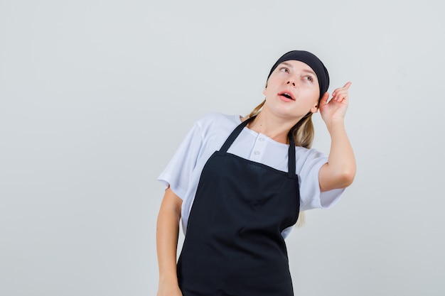 Foto gratuita joven camarera en uniforme y delantal inclinando la cabeza y apuntando hacia arriba