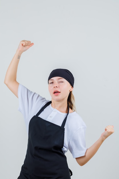 Foto gratuita joven camarera en uniforme y delantal mostrando gesto de ganador y mirando confiado