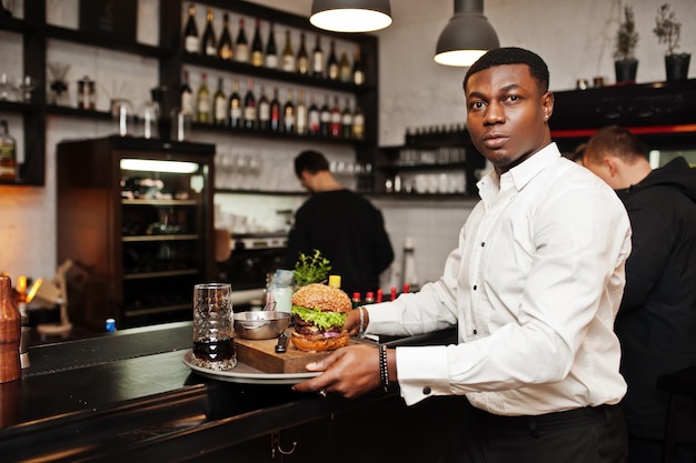 Joven camarero afroamericano sostiene bandeja con hamburguesa en el bar del restaurante