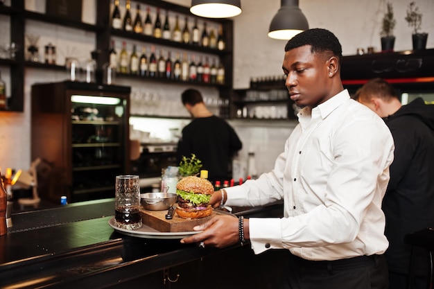 Joven camarero afroamericano sostiene bandeja con hamburguesa en el bar del restaurante