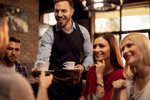 Joven camarero feliz dando café a sus clientes mientras les sirve en una cafetería